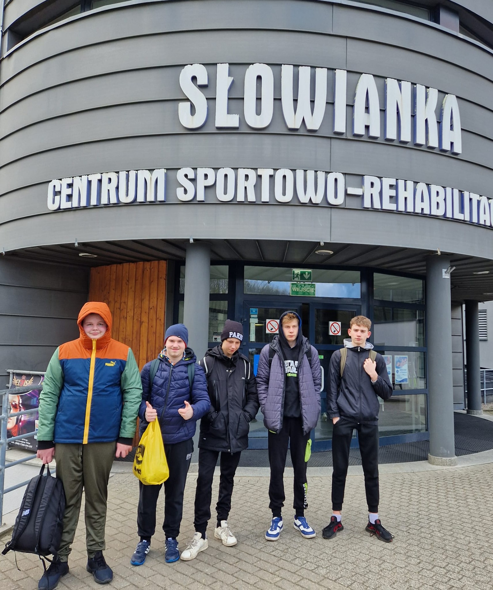 5 uczniów w wieku 15-20 lat stoi przed Centrum Sportowo - Rekreacyjnym Słowianka w Gorzowie. Są ubrani w ciepłe kurtki i czapki.