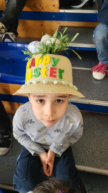 Chłopiec siedzący na trybunach. Patrzący w górę z obiektyw aparatu. Na głowie ma słomkowy kapelusz. Na nim napis Happy Easter z kolorowych liter. 