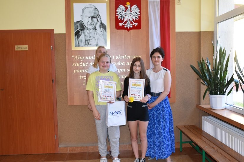 Paulina i Klaudia wraz z nauczycielką języka niemieckiego i wicedyrektor szkoły przed tablicą poświęconą patronowi szkoły Janowi Pawłowi II