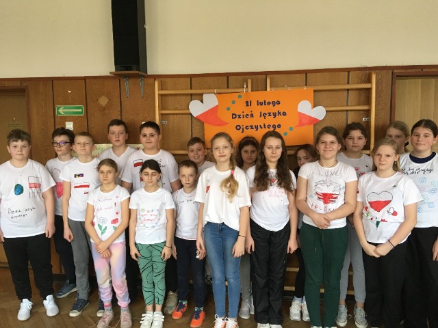 17 uczniów stoi ubranych w białe koszulki, na których narysowali symbole związane z Międzynarodowym Dniem Języka Ojczystego. Nad nimi wisi pomarańczowy plakat z napisem Międzynarodowy Dzień Języka Ojczystego.