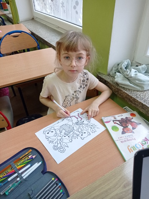 W ławce szkolnej siedzi dziewczynka w okularach, na ławce ma kolorowankę i podręcznik.