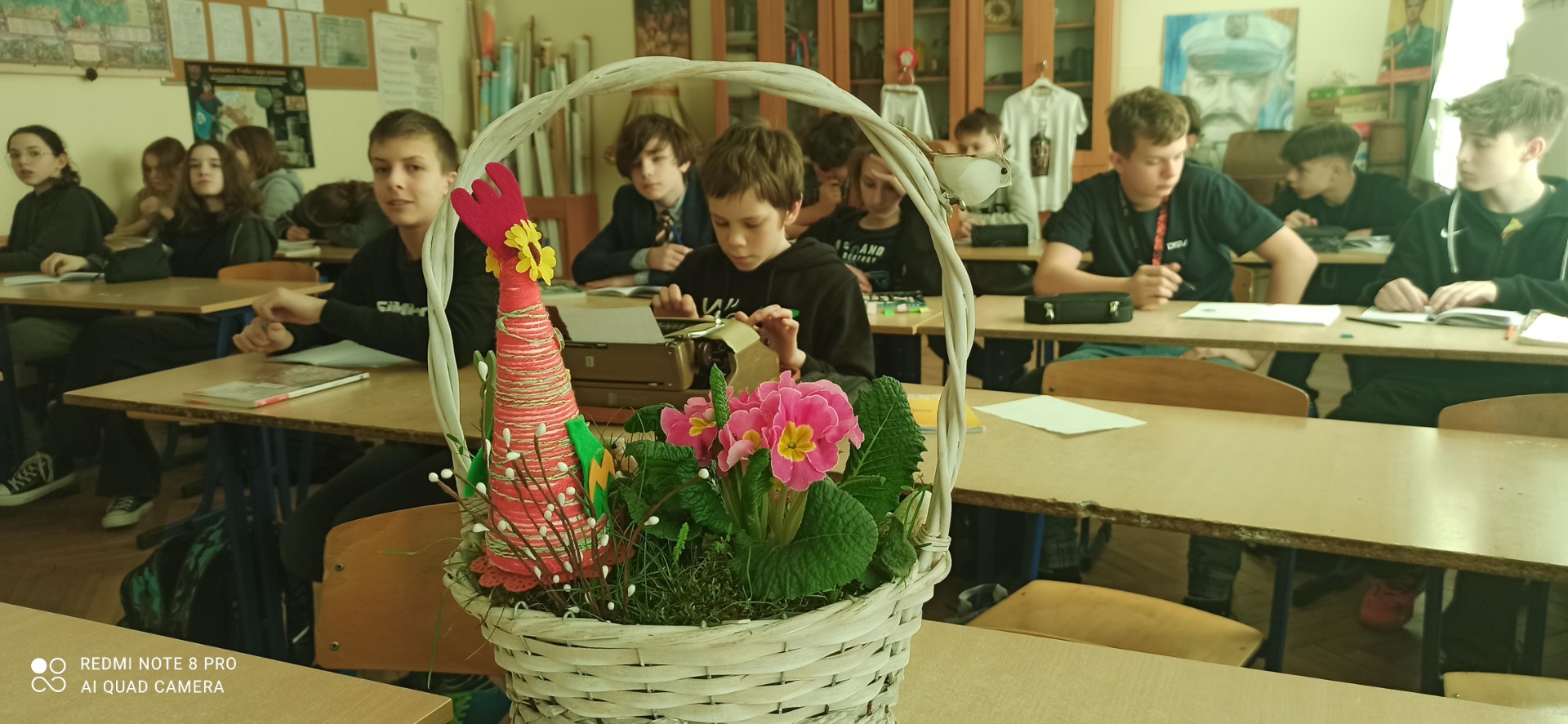 Na pierwszym planie widoczny koszyczek wielkanocny z kwiatkami i papierowym kurczakiem. W tle uczniowie kl. 8a siedzący przy stolikach