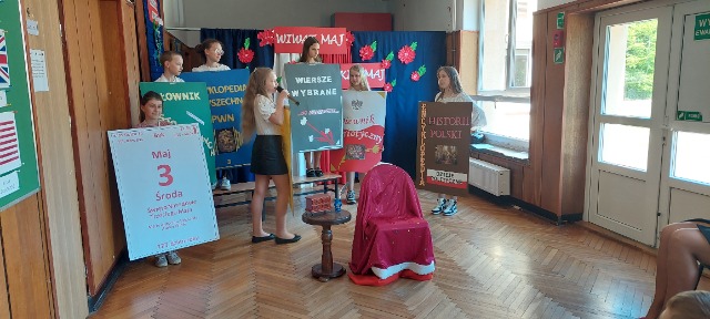 Na pierwszym planie stoi  krzesło przystrojone czerwona tkaniną i mały stolik. Dalej stoi sześć uczennic i jeden uczeń. Uczniowie trzymają okładki książek wykonane z dużego kartonu.