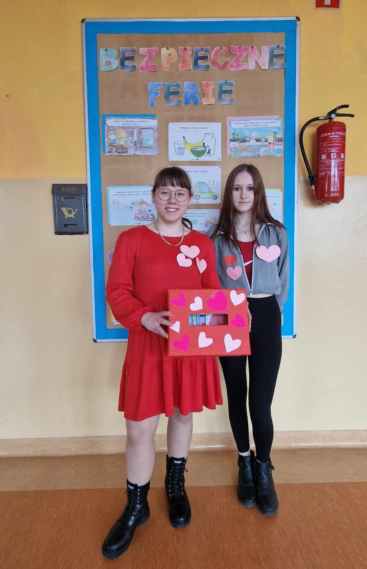 Uczennice - wolontariuszki ubrane w czerwone dodatki prezentują do zdjęcia ze skrzynką walentynkową