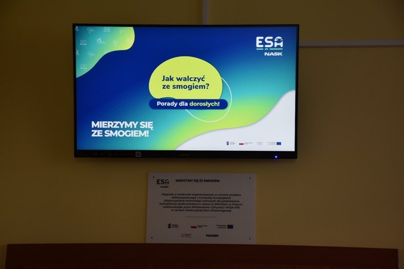 Monitor w wyświetlanymi informacjami o sposobach poprawiania jakości powietrza  i zamieszczona pod nim tabliczka z napisem ESA - Mierzymy się ze smogiem.