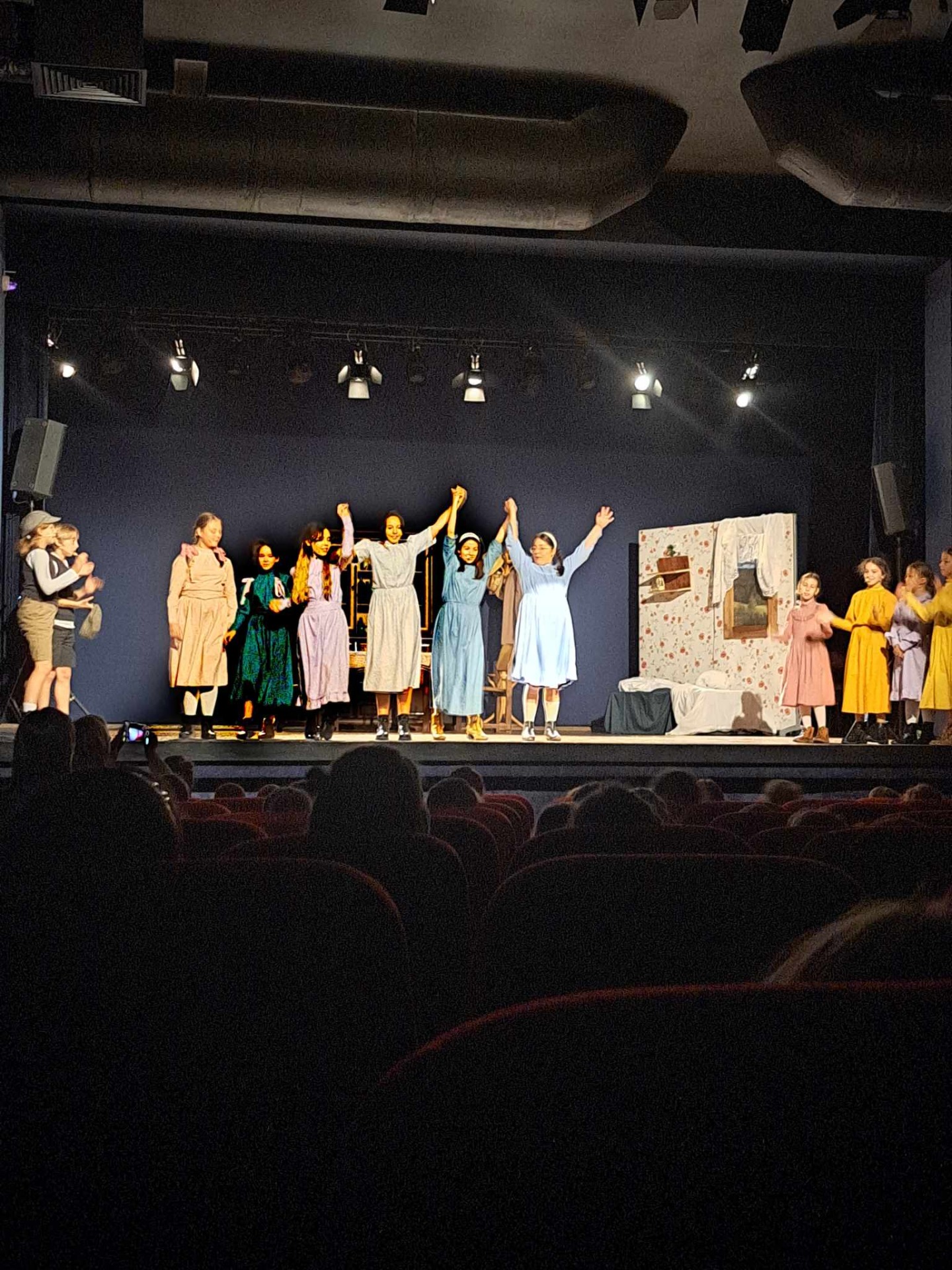 Na scenie stoi młodzież w stylowych strojach - aktorzy. Trzy dziewczynki podnoszą ręce do góry.