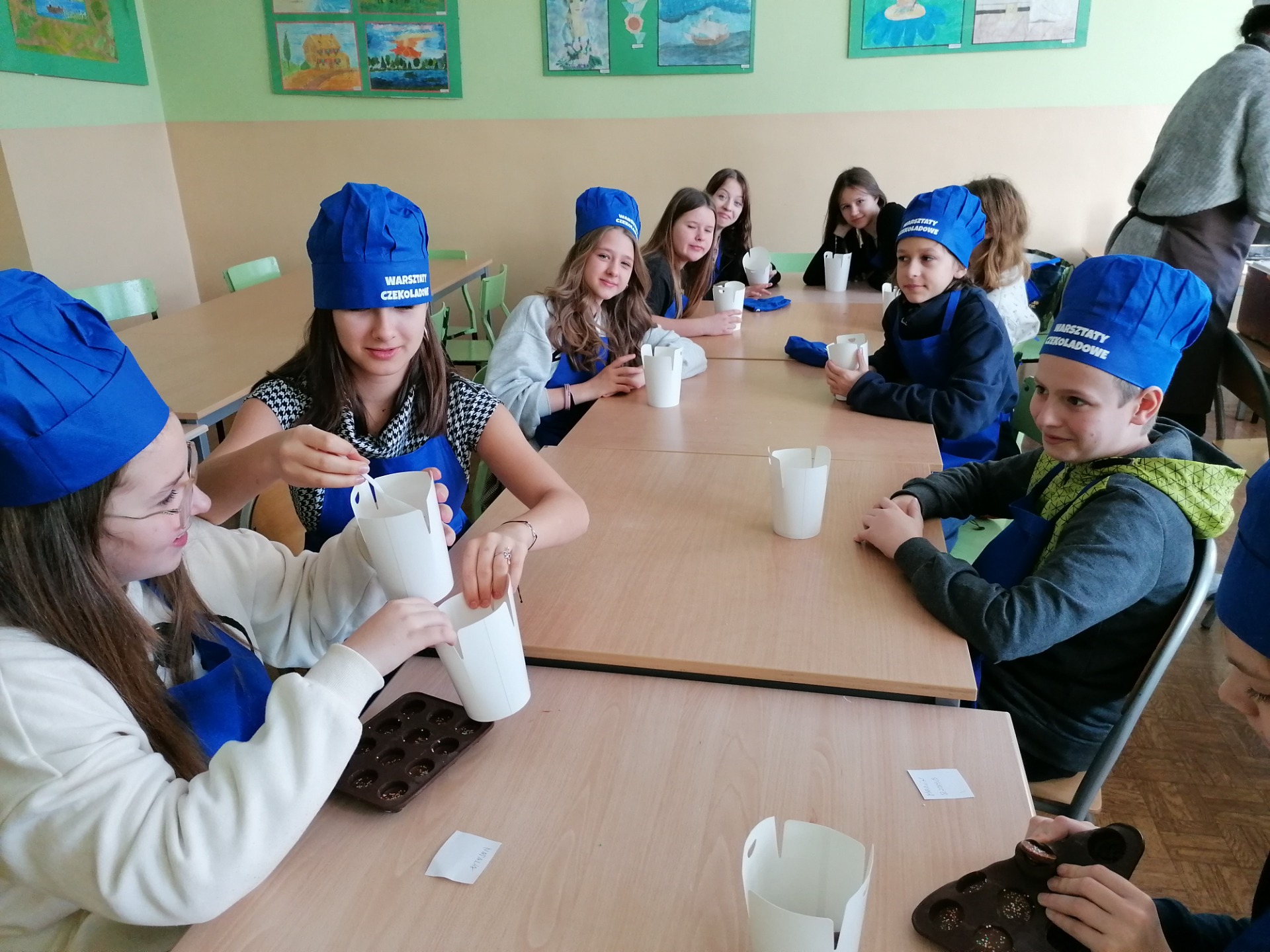 Grupa uczniów w niebieskich czepkach siedzi przy stole, przed nimi białe kubeczki
