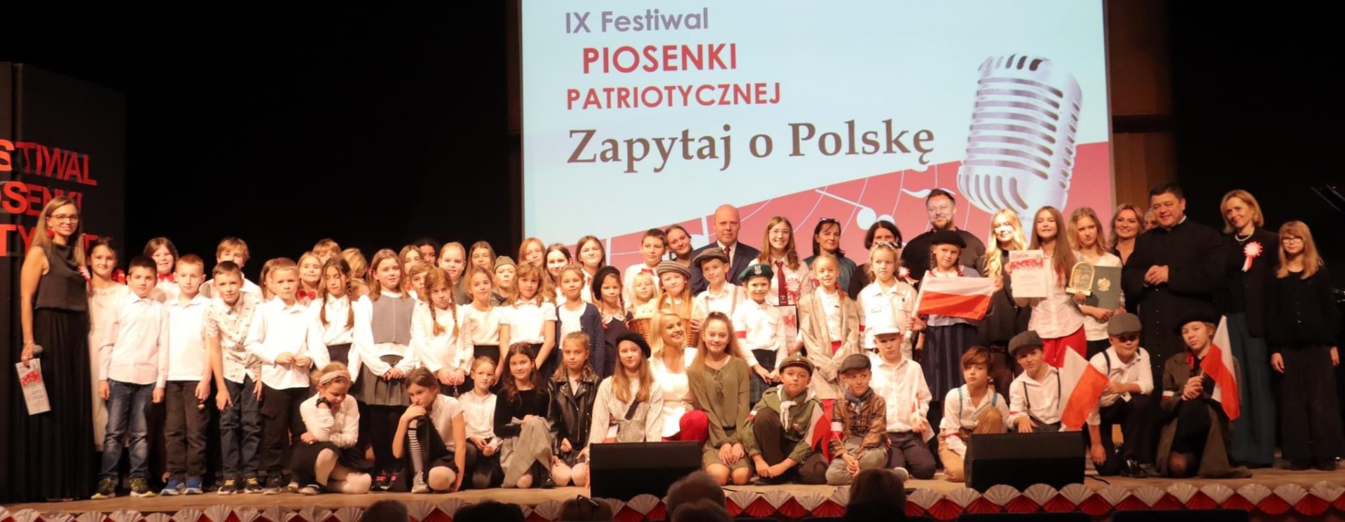 Dzieci na scenie w obecności nauczycieli biorący udział w Festiwalu Pieśni Patriotycznej