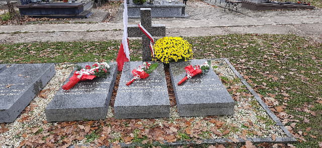 Trzy płyty nagrobkowe, na których leżą wiązanki kwiatów przybrane w biało-czerwone barwy. Za nagrobkami krzyż, obok flaga biało-czerwona  i chryzantemy.