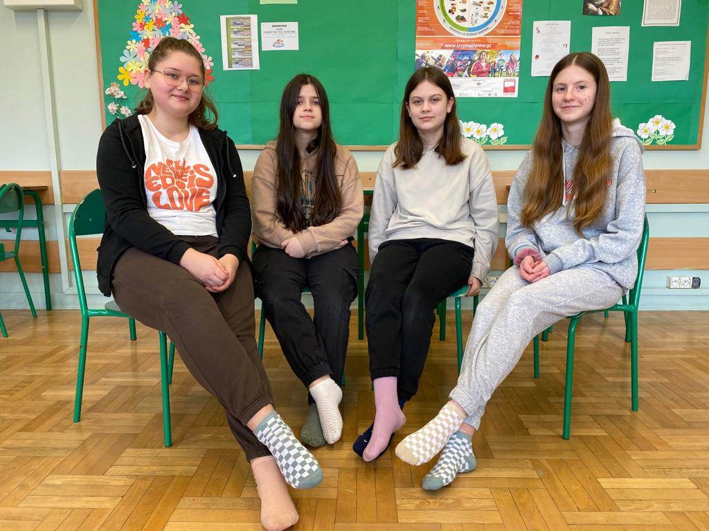Na zdjęciu, w klasie, na zielonych krzesłach siedzi cztery dziewczynki klasy siódmej. Wszystkie mają zdjęte buty i pokazują stopy ubrane w skarpetki nie do pary. Za nimi w tle  klasowa gazetka.