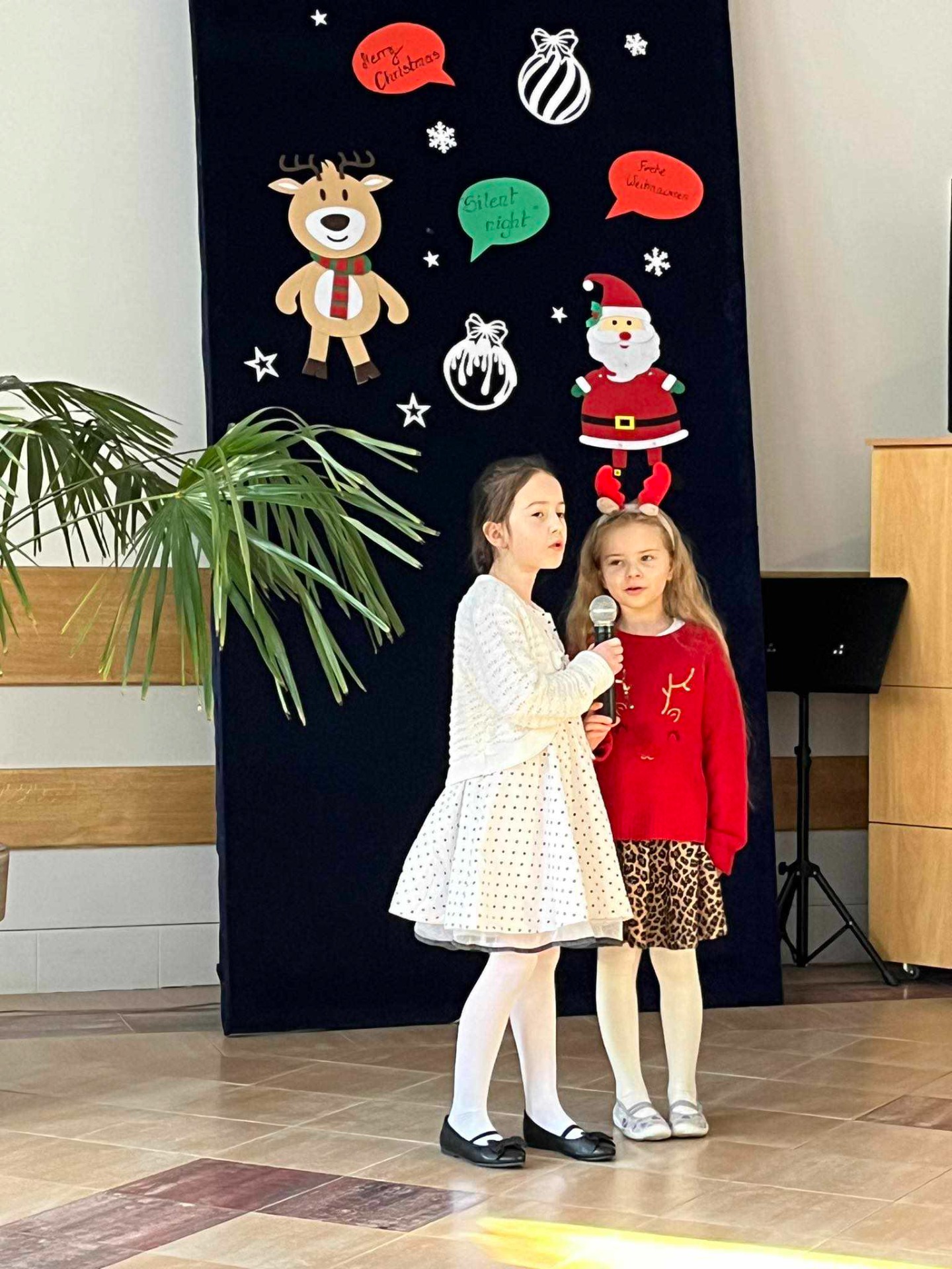 Na tle świątecznej dekoracji stoją dwie dziewczynki ubrane na czerwono i biało, śpiewają do mikrofonu.