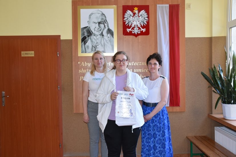 Amelka wraz z nauczycielką języka niemieckiego i wicedyrektor szkoły przed tablicą poświęconą Janowi Pawłowi II