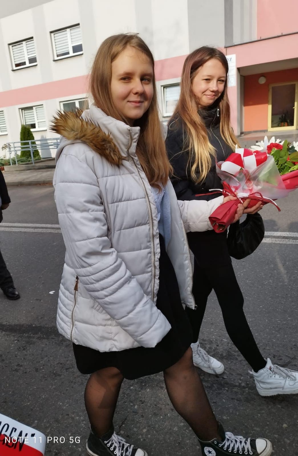 Dwie dziewczyny trzymają wiązankę biało czerwonych kwiatów. Jedna ubrana w biała kurtkę druga w czarną.