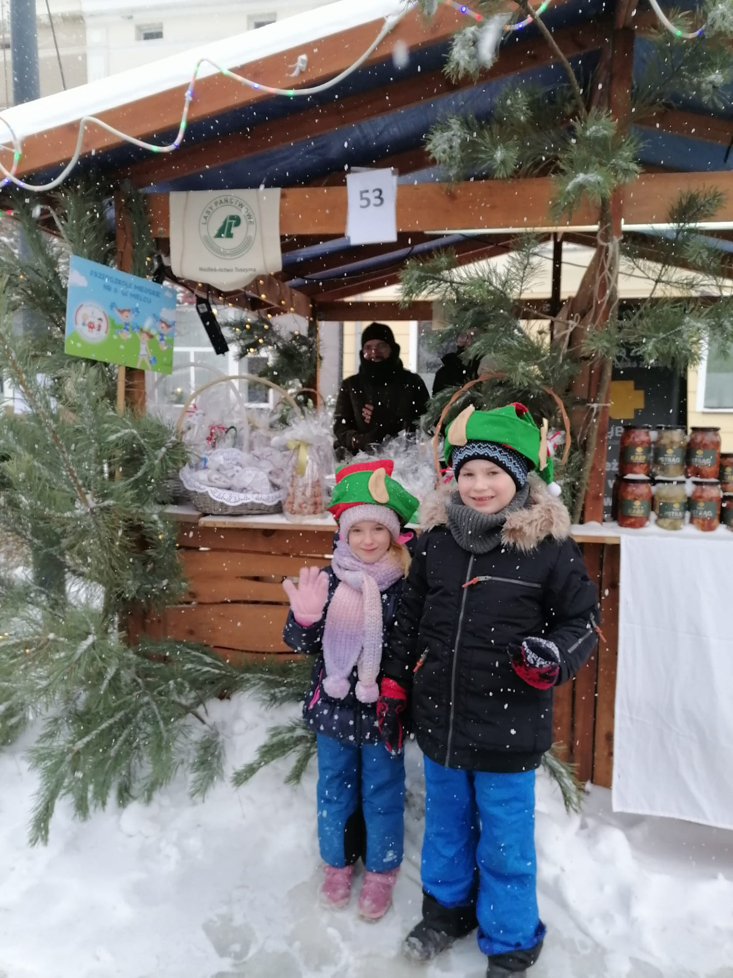 Na pierwszym planie pośrodku zdjęcia stoi dwójka dzieci chłopiec i dziewczynka w elfich czapkach na głowie. Za nimi znajduje się drewniane ne stoisko ozdobione zielonymi gałązkami choinki.