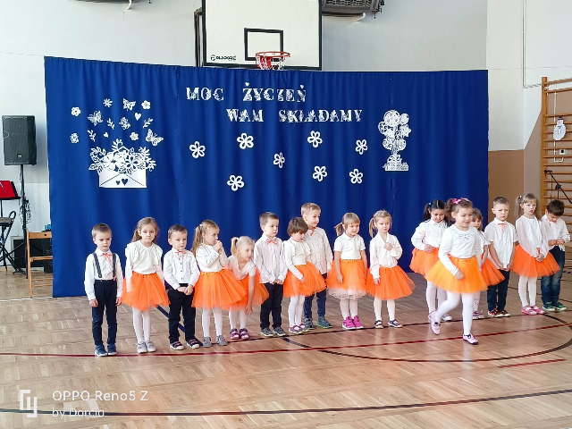 Występ przedszkolaków 4-5 latków na tle niebiesko białej dekoracji na dzień Babci i Dziadka.