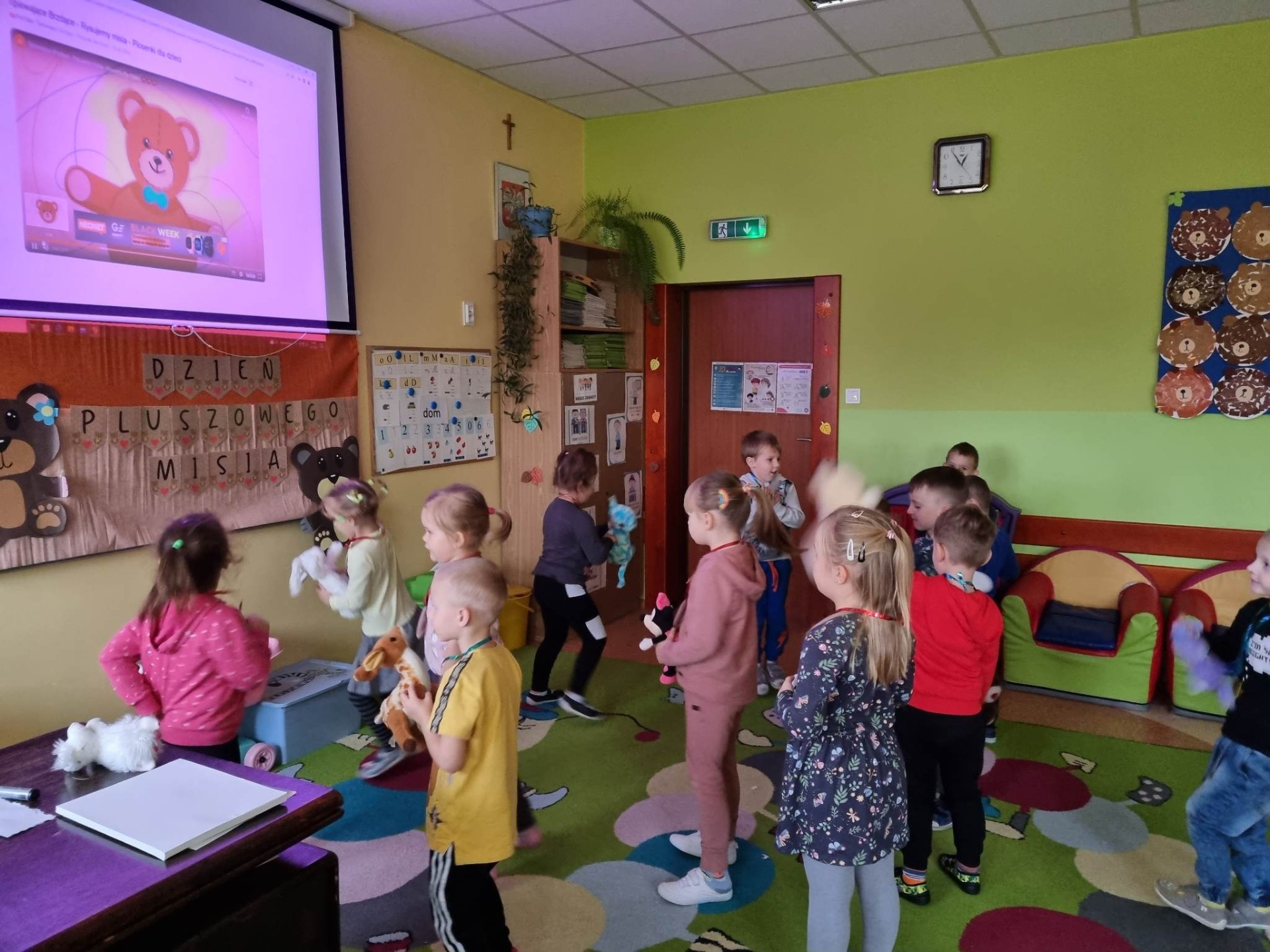 Przedszkolaki tańczą w sali przedszkolnej w rytm misiowych piosenek wyświetlanych na ekranie